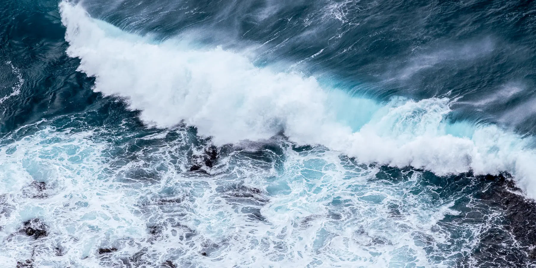 A turquoise wave breaking over dark rocks, photographed by Adam Grimm